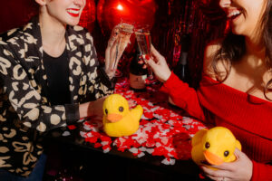 Two women drinking prosecco over a confetti covered table with stuffed duck toys 